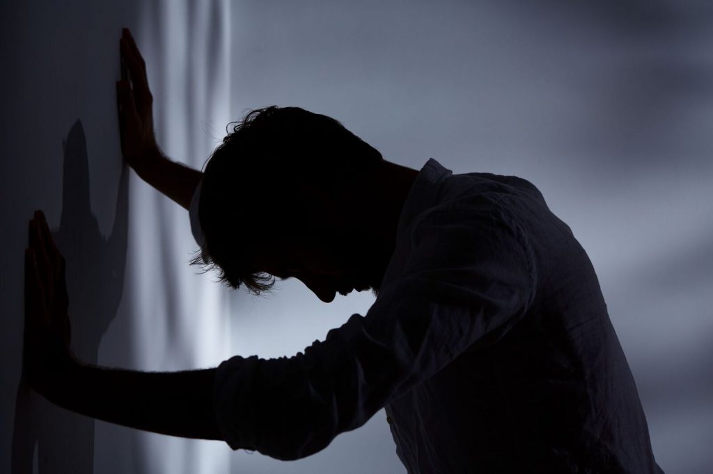 Man struggling with mental health with palms against the wall and his head down