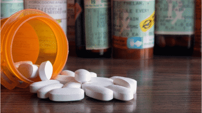 Opioid pills spilling out of an orange prescription bottle in front of various other prescription bottles